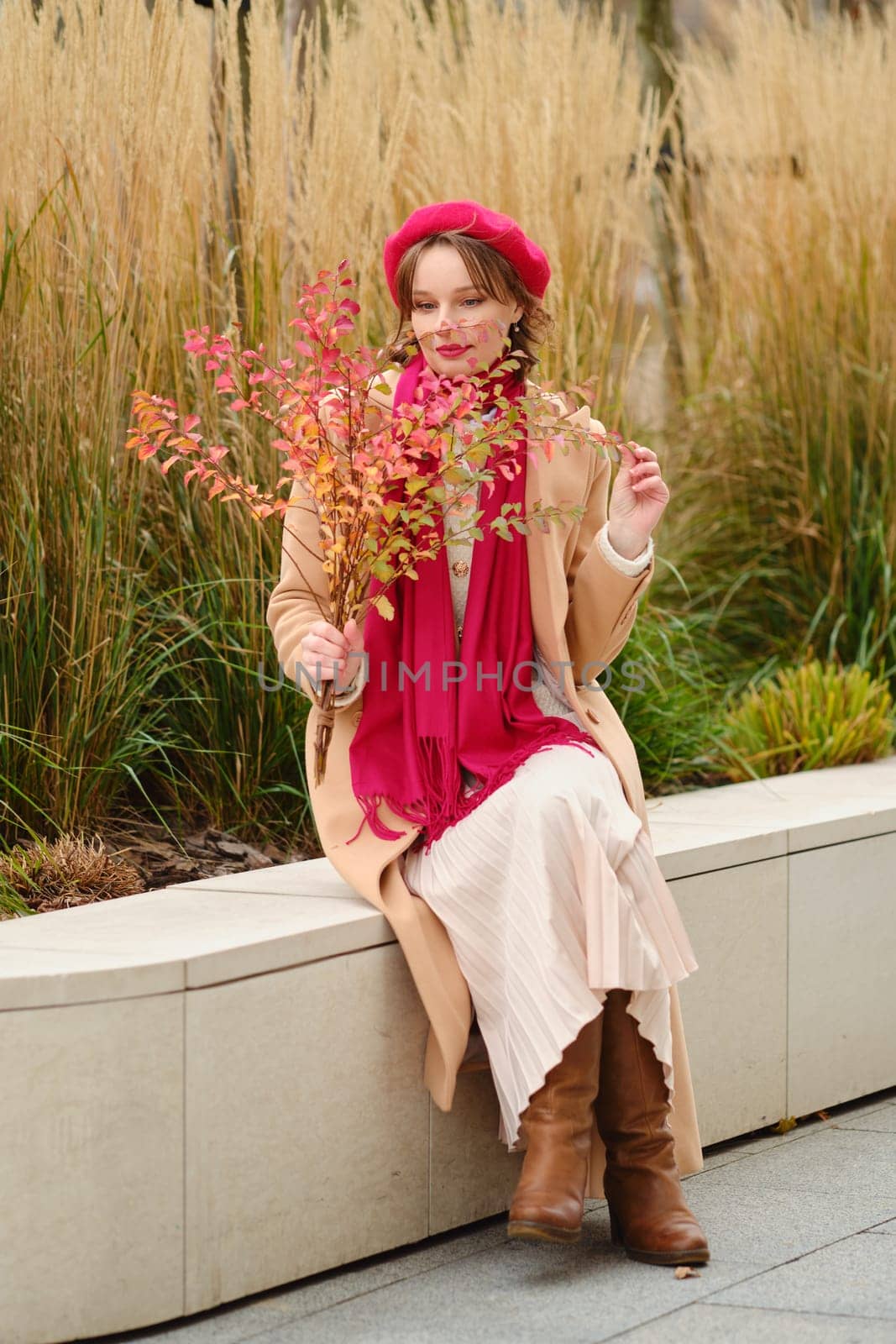 A woman wearing a red beret and a stylish coat enjoys a tranquil afternoon in the park, holding colorful autumn leaves. The setting features tall grasses and greenery, creating a serene atmosphere.