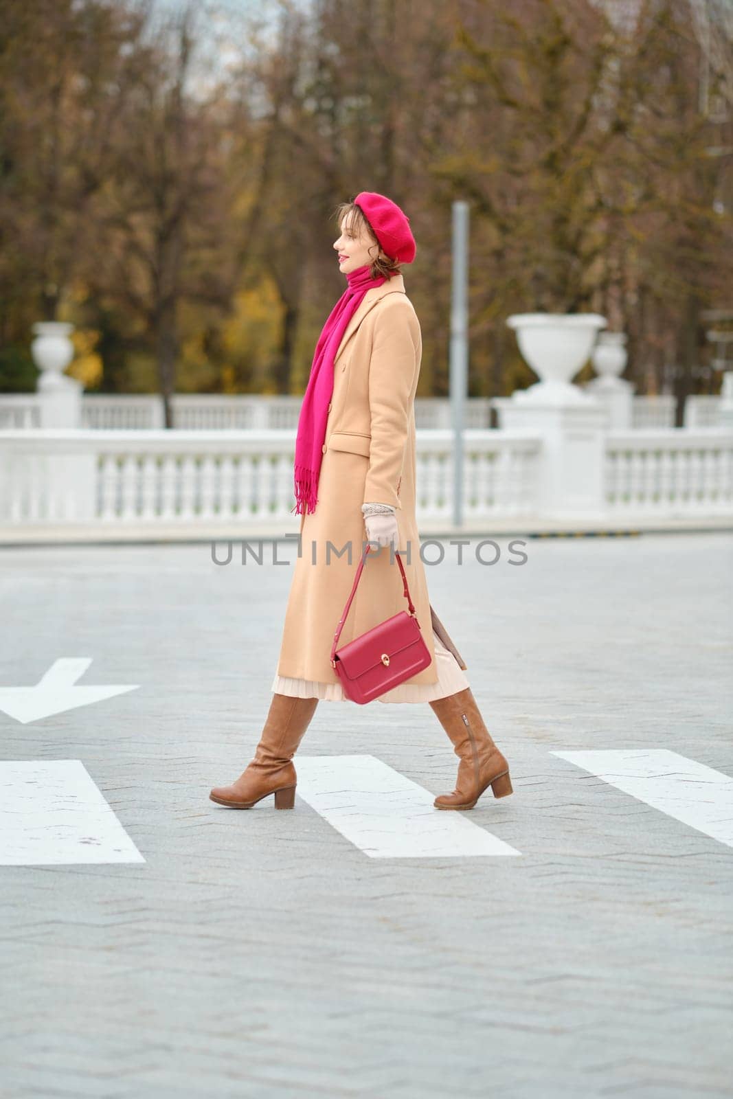 A woman wearing a beige coat, red beret, and a bright scarf walks across a crosswalk in a scenic park setting, showcasing elegant fashion and autumn scenery while carrying a stylish handbag.