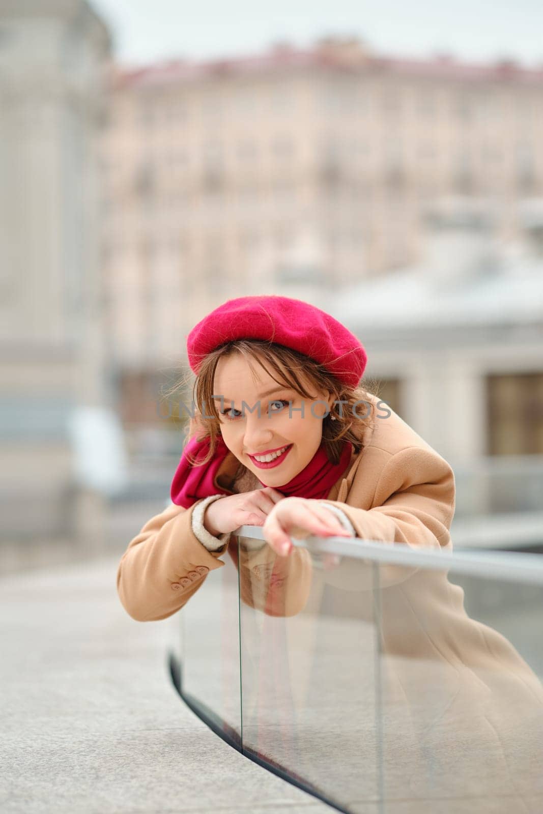 In a lively city backdrop, a cheerful young woman in a red beret leans playfully on a glass railing, enjoying her surroundings in the soft light of day, radiating joy and style.