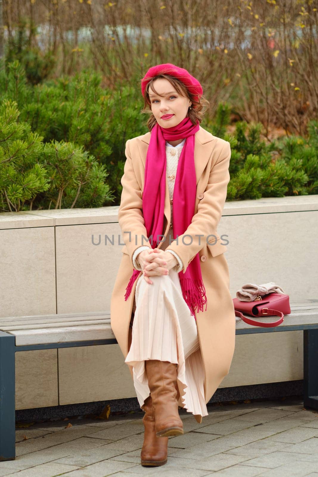 A woman sits comfortably on a park bench, dressed warmly in a beige coat, pink scarf, and tall brown boots while enjoying the autumn ambiance surrounded by greenery and fallen leaves.