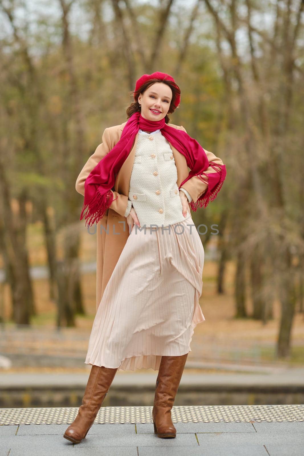 A fashionable woman stands playfully in a warm, stylish outfit featuring a scarf and long skirt. The autumn park setting adds to her charm as trees with colourful foliage surround her.