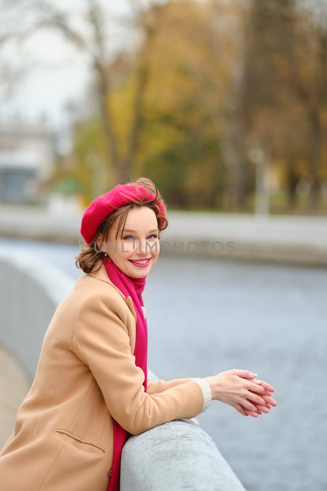 A joyful woman wears a red beret and beige coat while resting her hands on a concrete railing by the river.