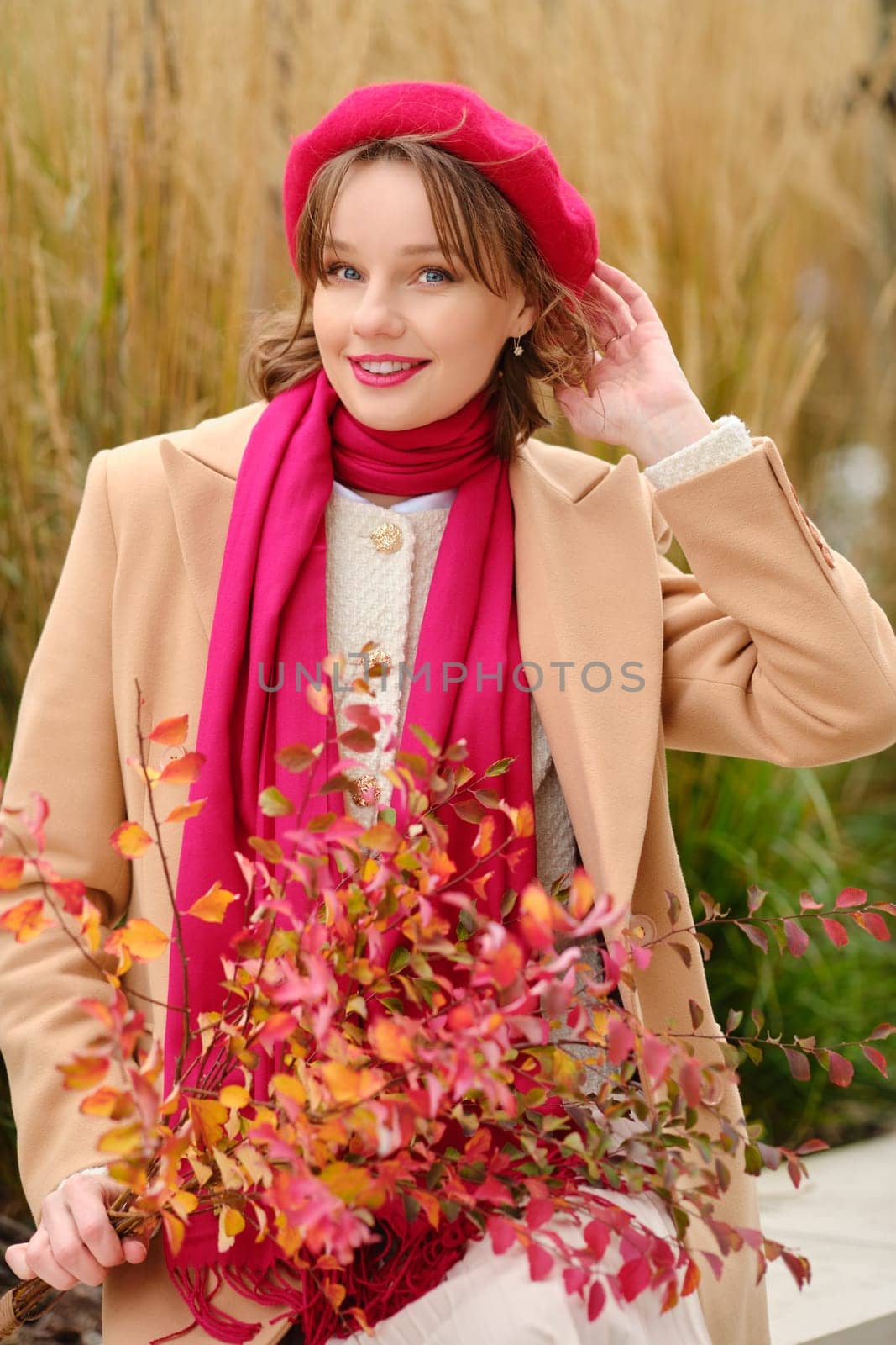 A woman dressed in a beige coat and pink scarf playfully poses outdoors. She holds a bouquet of vibrant autumn leaves, showcasing the beauty of the season against a natural backdrop.