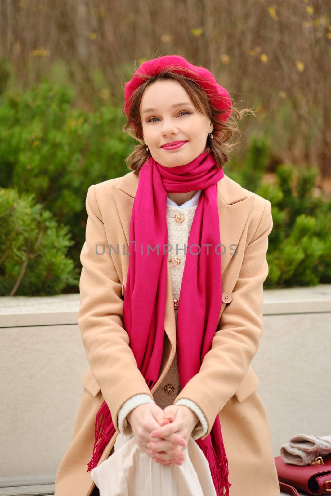 A young woman sits outdoors, dressed in a stylish coat and a vibrant scarf. She gazes happily at the camera, surrounded by greenery on a sunny autumn day, exuding warmth and charm.