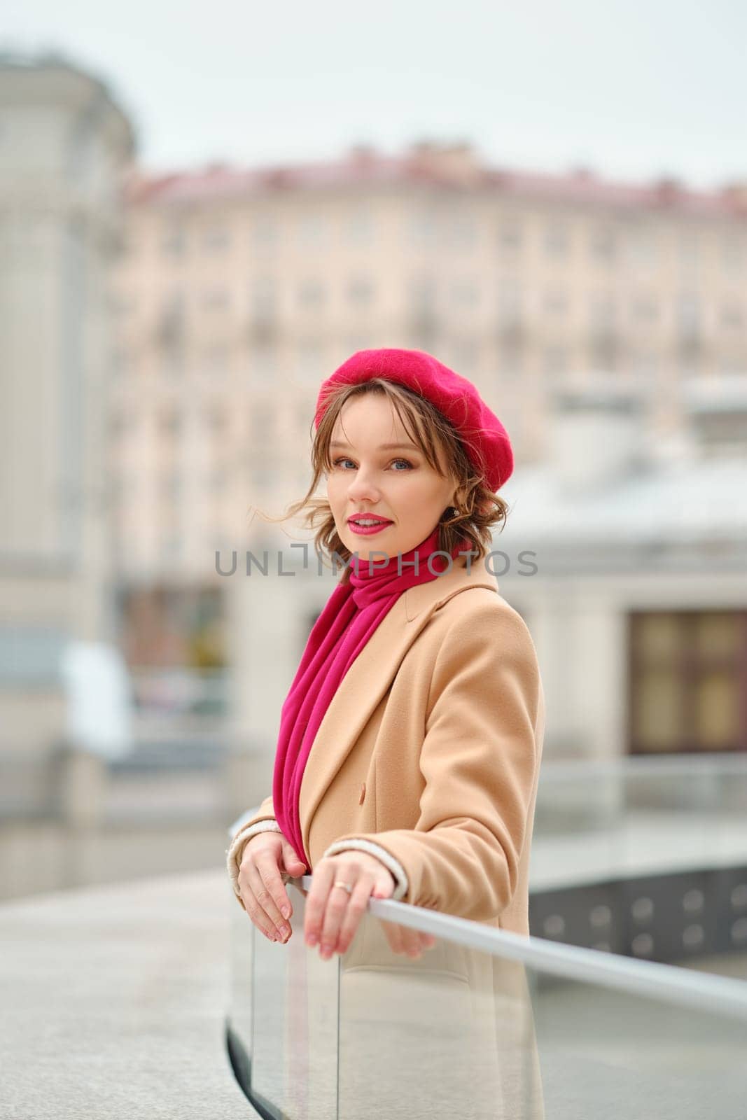 A stylish woman stands by a railing with a soft smile, wearing a red beret and matching scarf, against an urban backdrop.
