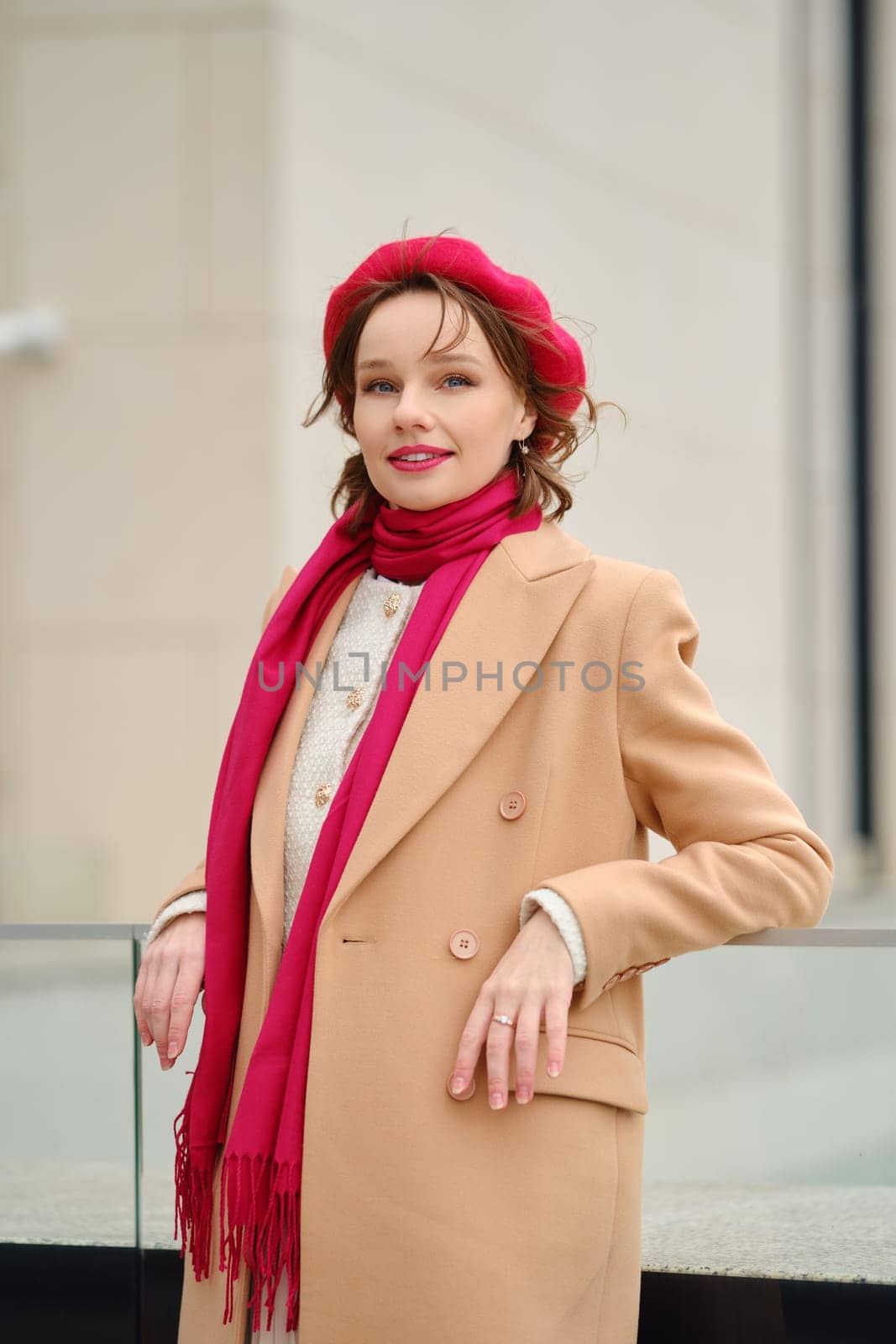 A fashionable woman stands outdoors wearing a stylish tan coat complemented by a vibrant red beret and scarf. Her confident pose showcases modern elegance on a cool day in an urban setting.