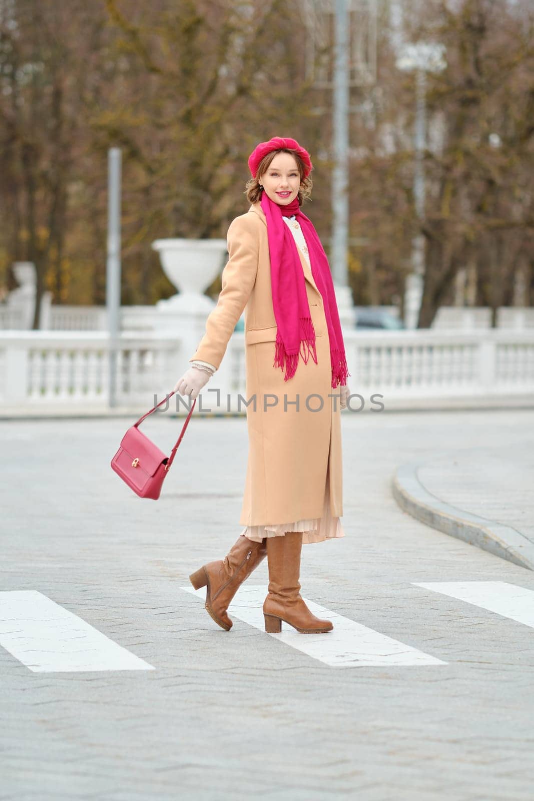 A fashionable woman is crossing the street in the city, wearing a beige coat, pink scarf, and red beret. She holds a bright pink handbag and walks confidently on the pedestrian crossing in autumn.