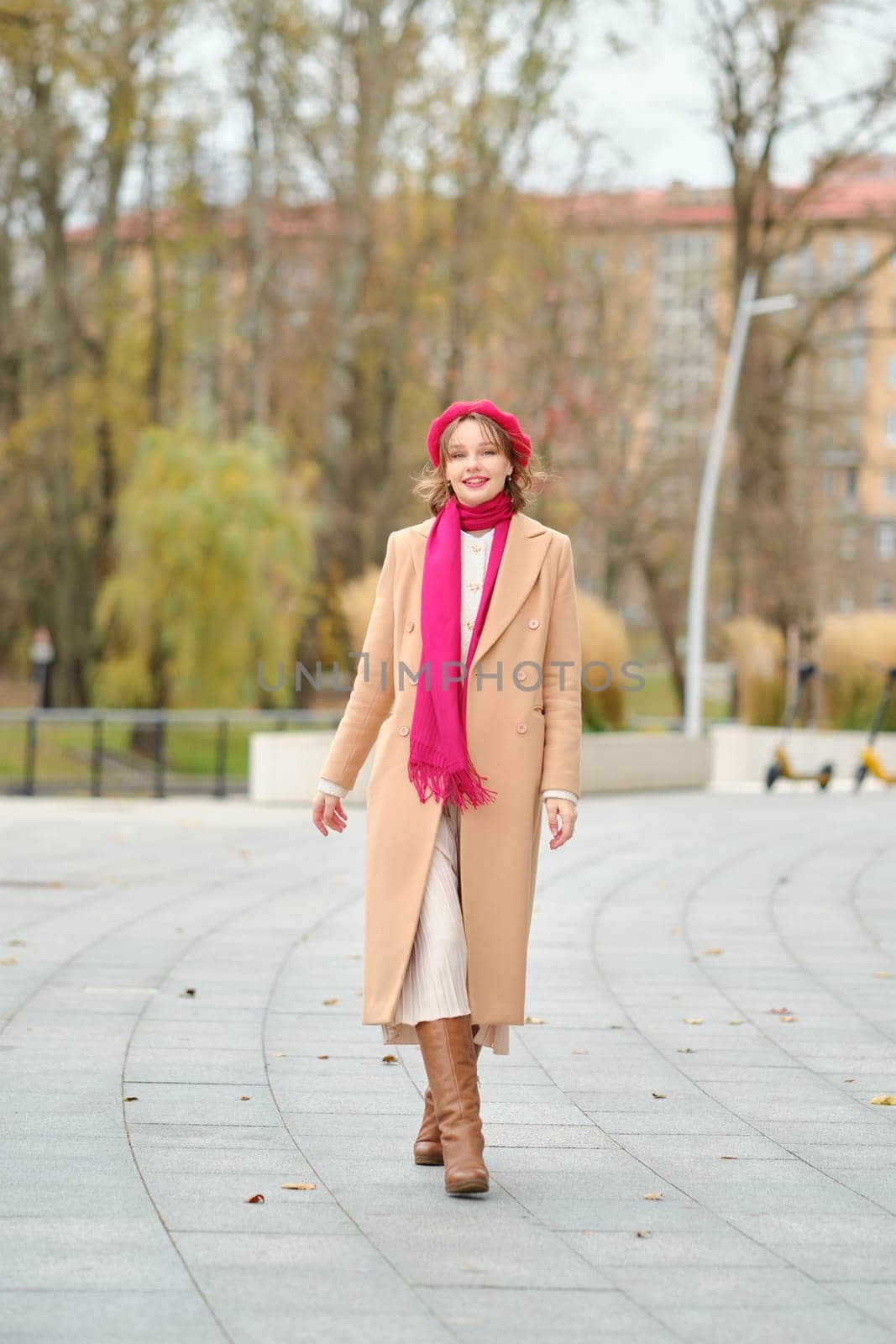 A woman walks through a picturesque park during autumn, dressed in a beige coat, scarf, and stylish boots. Her joyful expression complements the vibrant fall colors surrounding her.