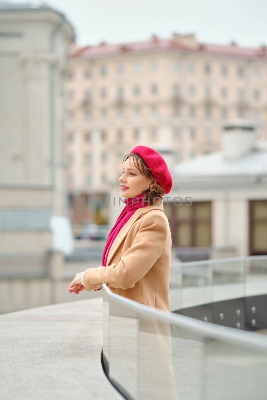 A woman wearing a red beret and matching scarf leans against a glass railing, gazing thoughtfully at the buildings in the background. The overcast sky adds a chilly atmosphere to the scene.