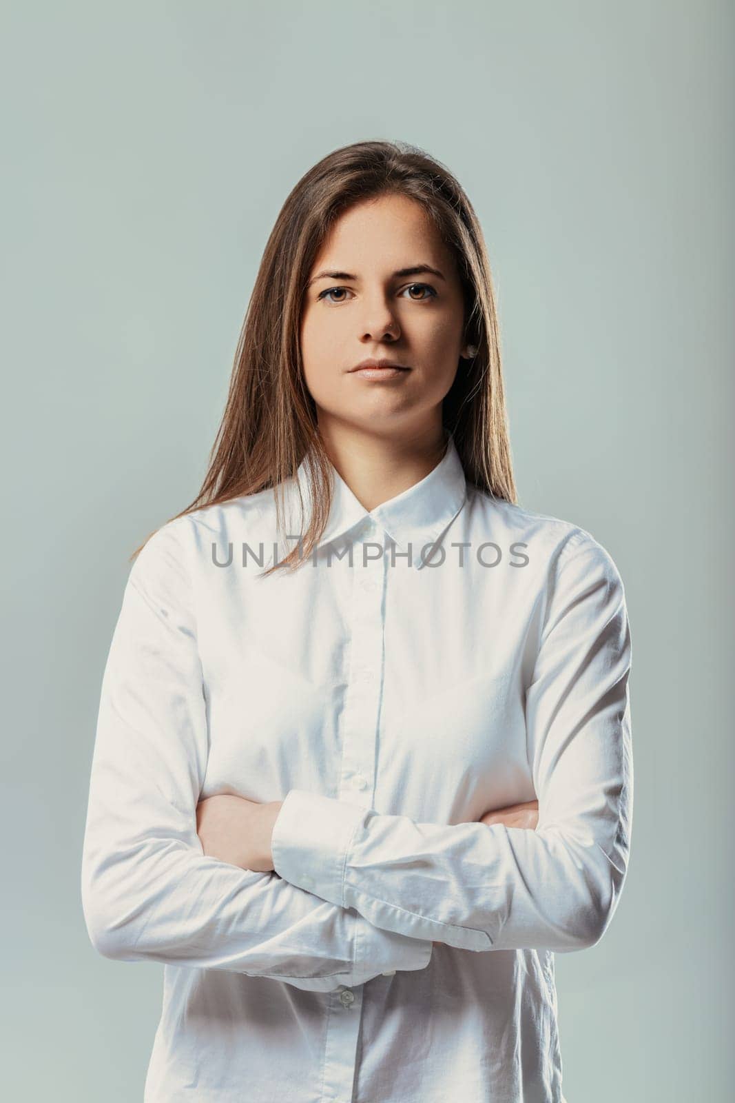 Studio portrait featuring a young businesswoman with arms crossed, exuding seriousness and confidence while showcasing determination and professionalism in a formal white shirt