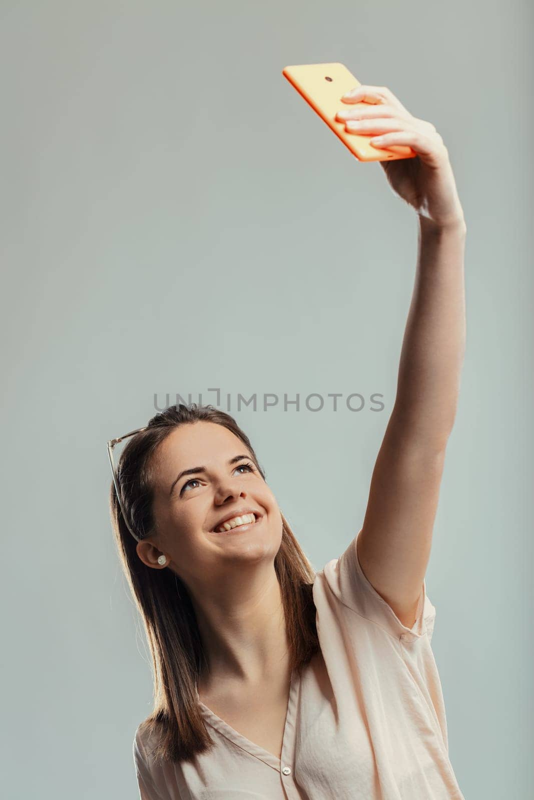 Cheerful young woman holding her smartphone high, capturing a selfie while looking up and smiling joyfully, radiating positivity and happiness against a gray studio backdrop