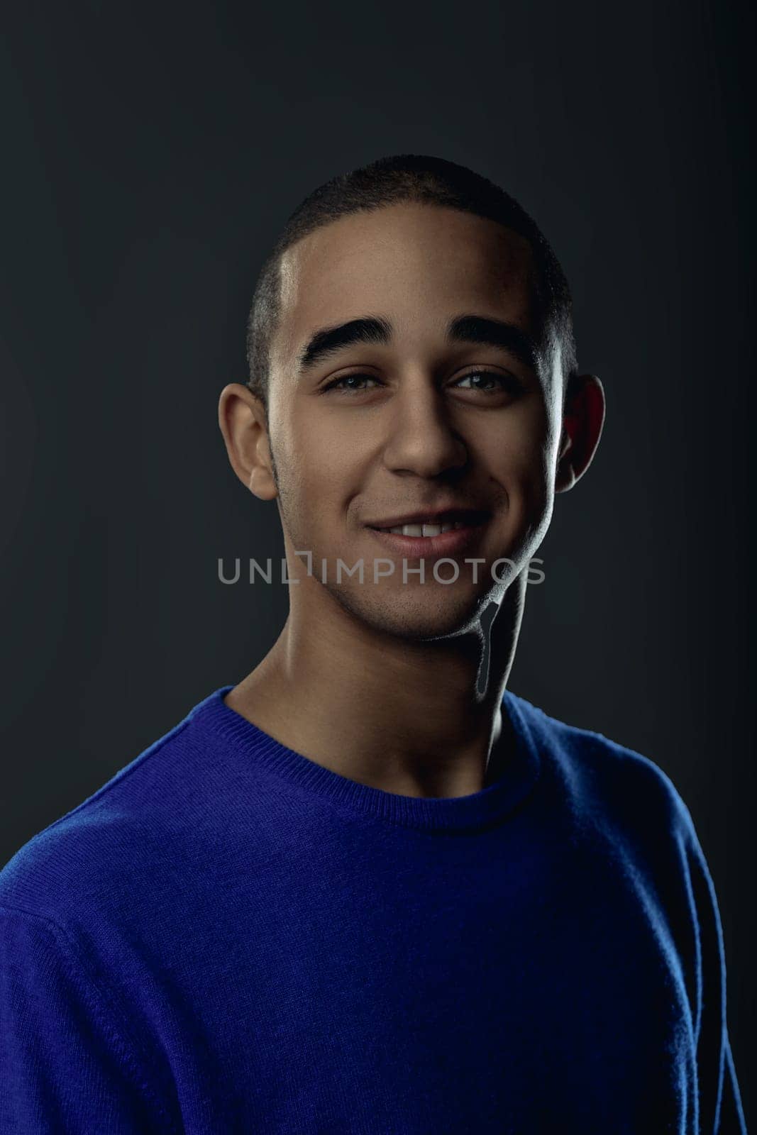 Portrait of a cheerful young man wearing a blue sweater, smiling and looking at the camera against a dark background. Portrait