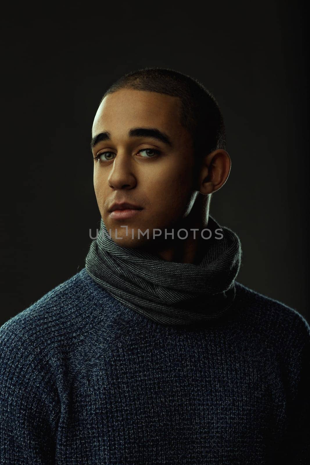 Portrait of a serious young man wearing a winter sweater and scarf, looking intensely at the camera against a dark background. Portrait