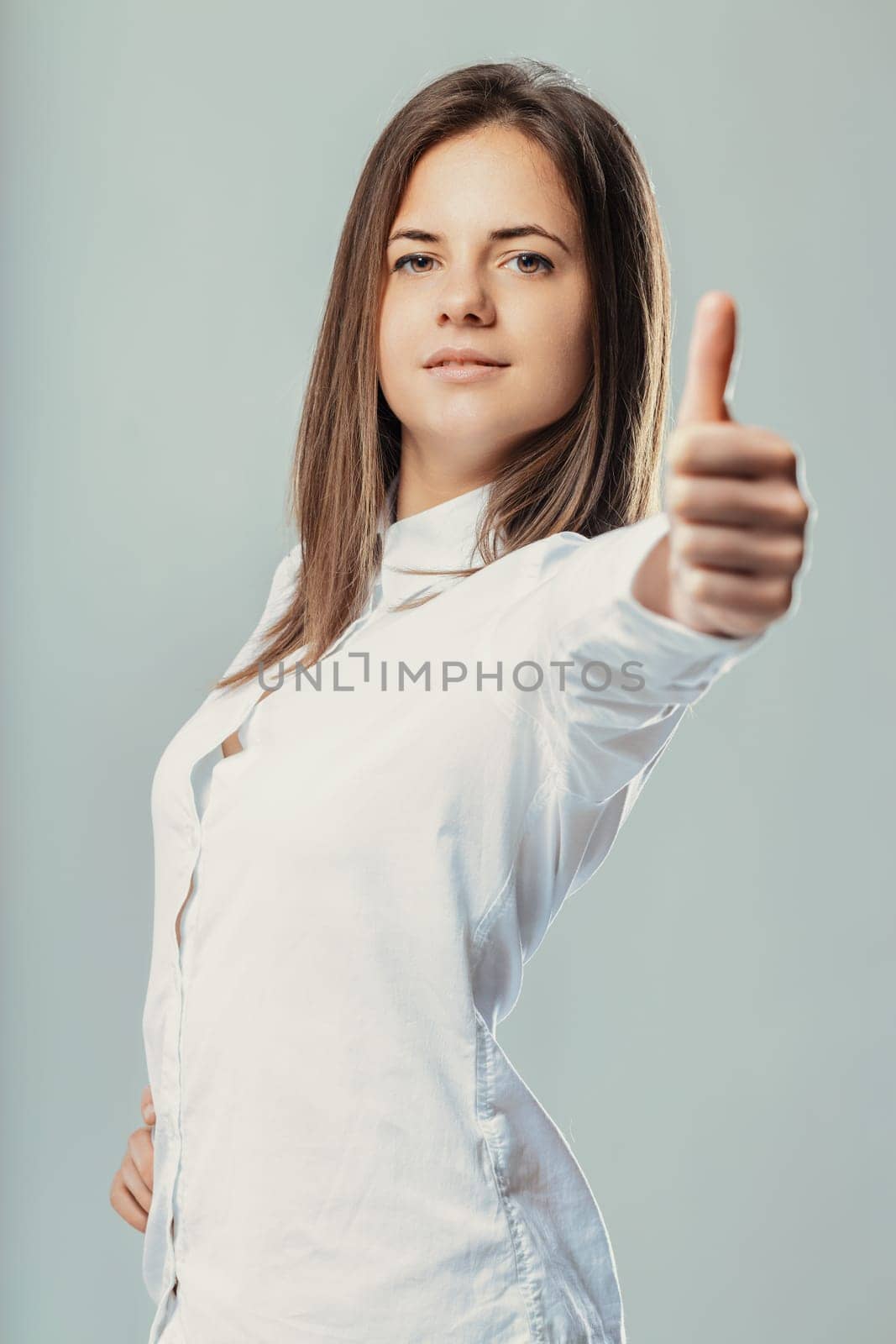 Young businesswoman smiling and showing a thumbs up while looking confidently at the camera, dressed in a white shirt against a light gray background, radiating positivity and empowerment