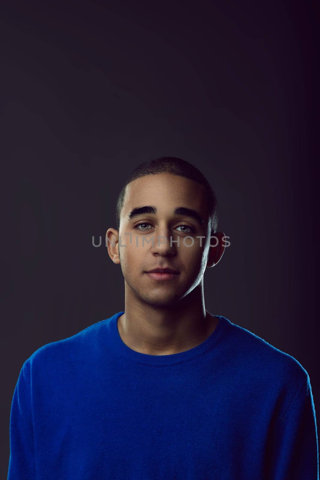 Portrait of a young man wearing a blue sweater, posing confidently while gazing directly at the camera against a dark background. Portrait