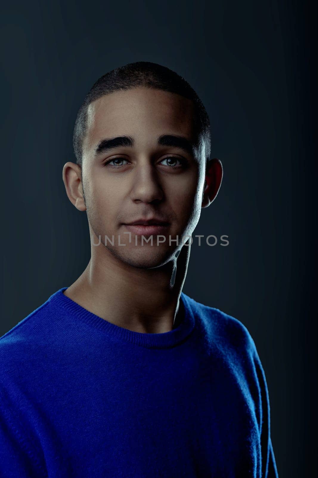 Portrait of a confident young man with short hair, wearing a blue sweater, posing against a dark studio backdrop. Portrait
