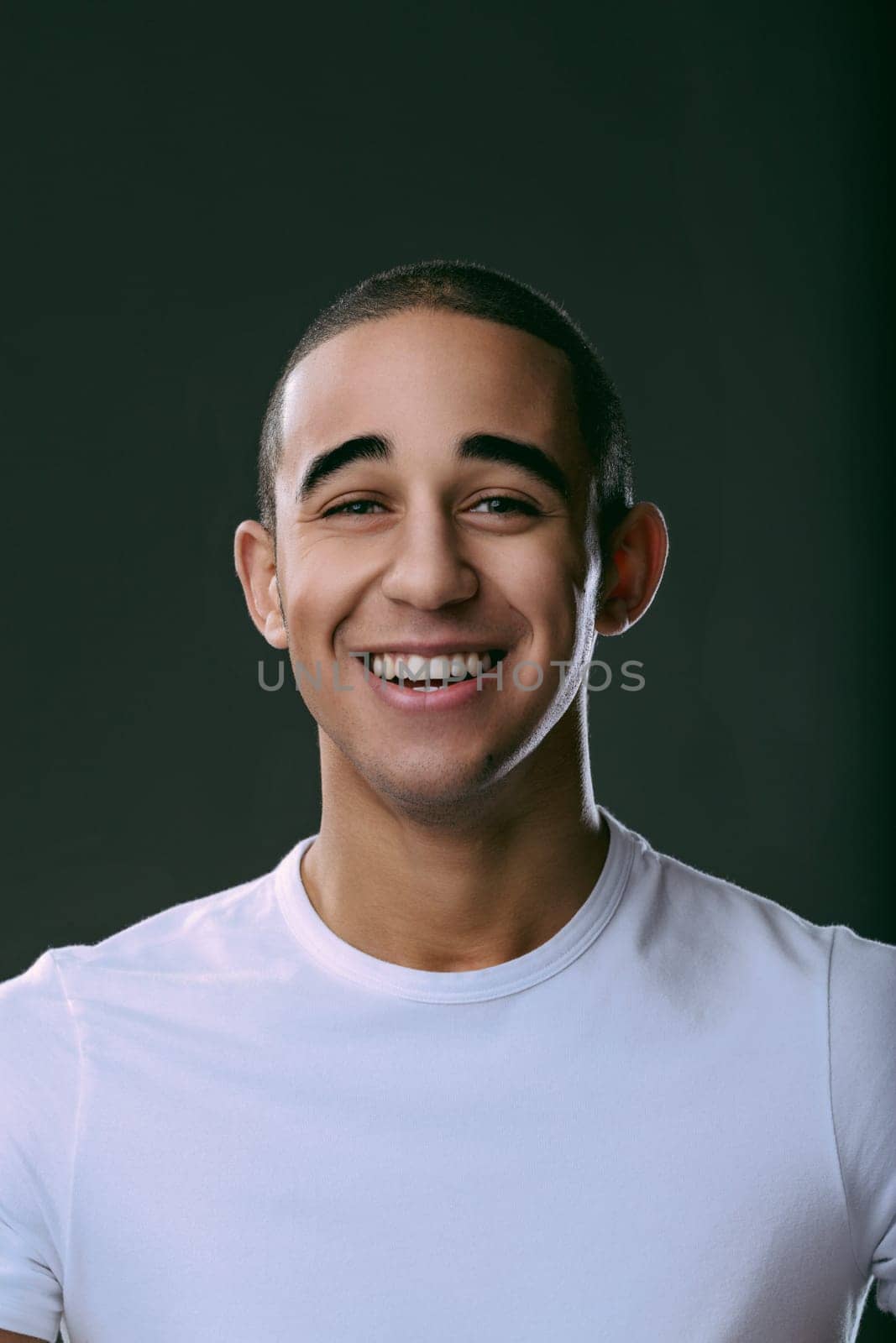 Cheerful young man smiling broadly, showcasing his teeth against a dark background, radiating happiness and confidence in a studio setting. Portrait