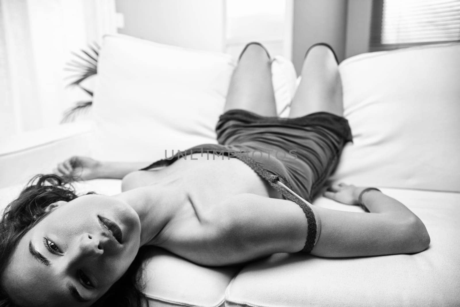 Young woman wearing a short dress lies on a white couch, gazing at the camera with a relaxed expression in a striking black and white photograph, exuding calm and elegance