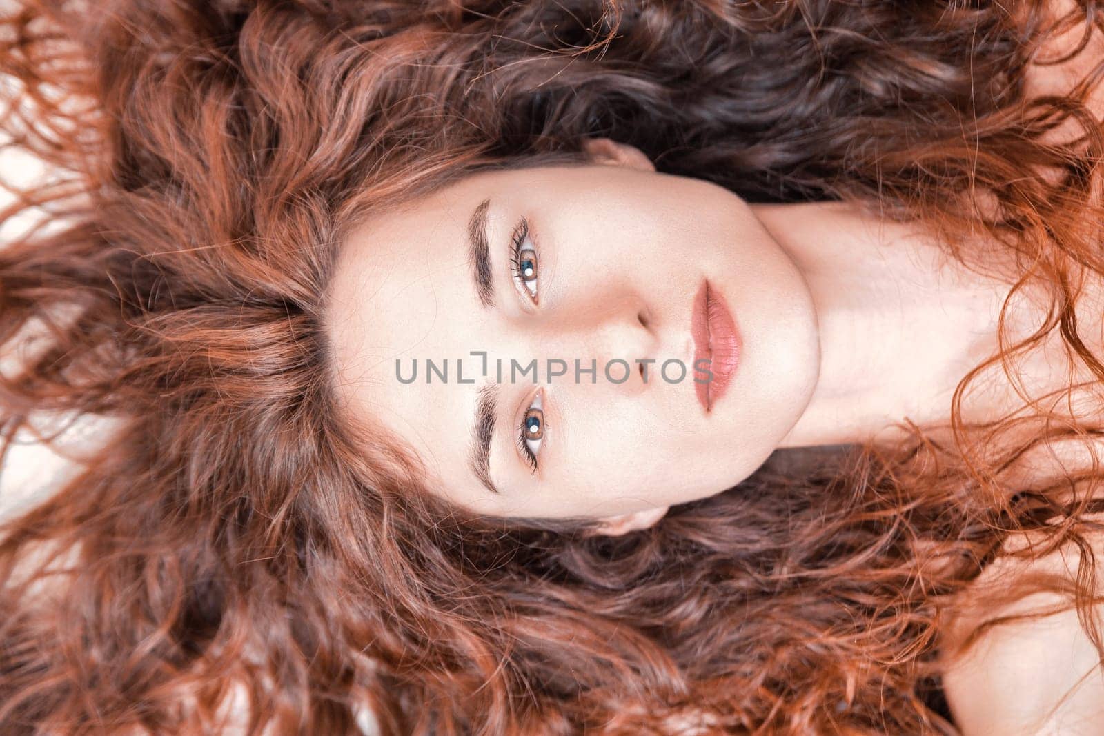 Close-up studio portrait featuring a stunning young woman with long, curly red hair, lying down and gazing at the camera with a serene and peaceful expression