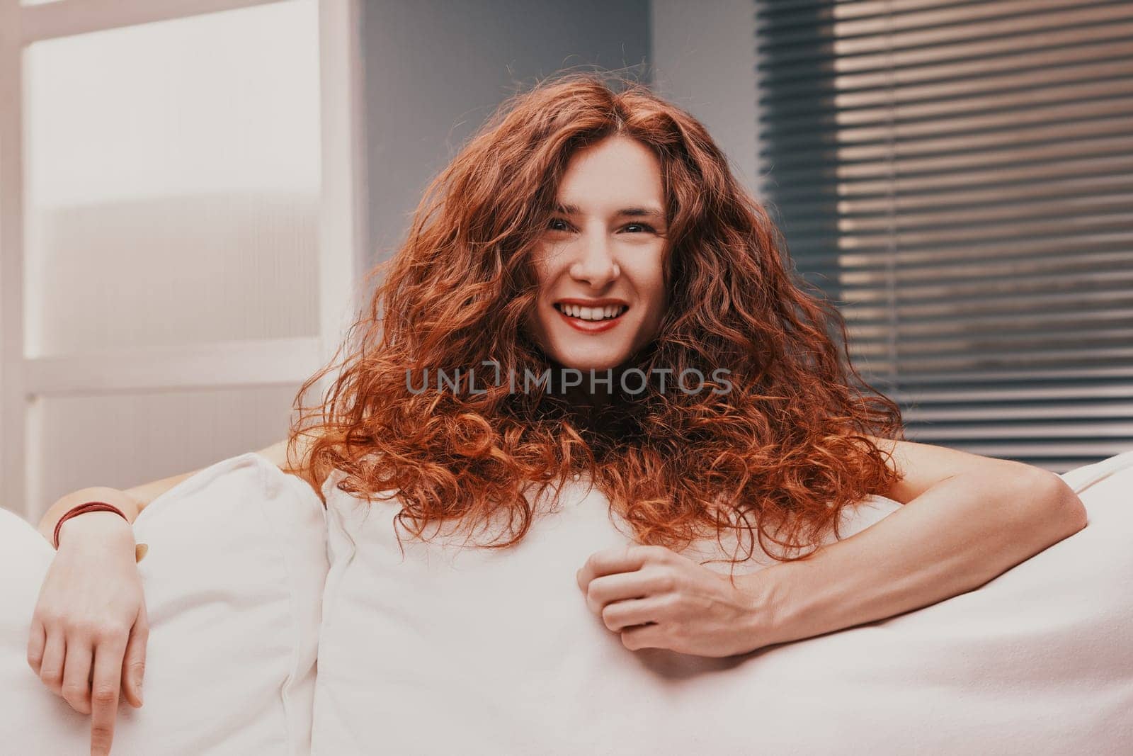 Young cheerful woman with long, curly red hair is smiling and relaxing on a comfortable white sofa in a cozy living room, enjoying a moment of peace and happiness