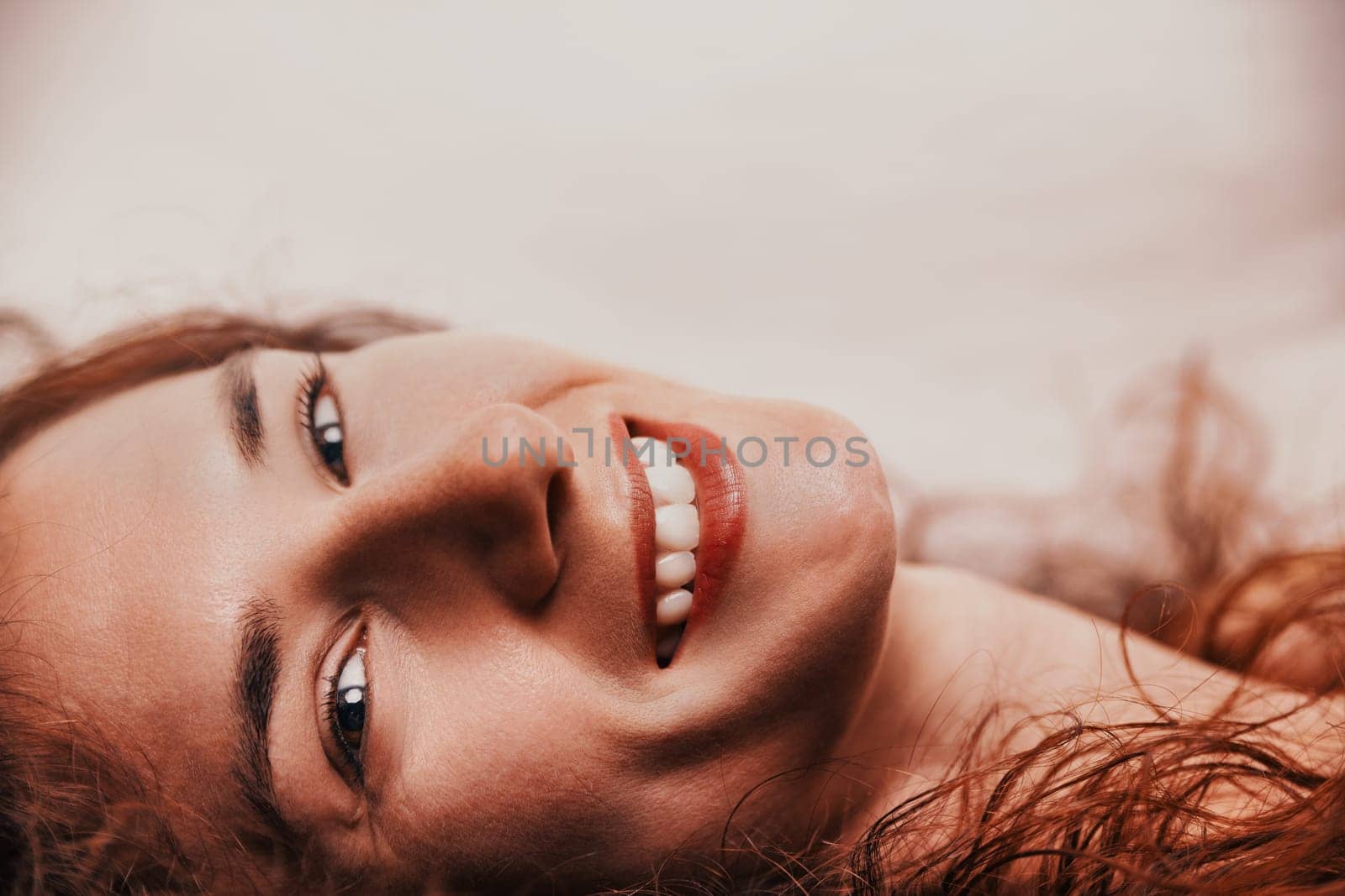 Close-up portrait featuring a cheerful young woman with long red hair lying down and smiling brightly, showcasing her teeth and exuding a sense of joy and contentment