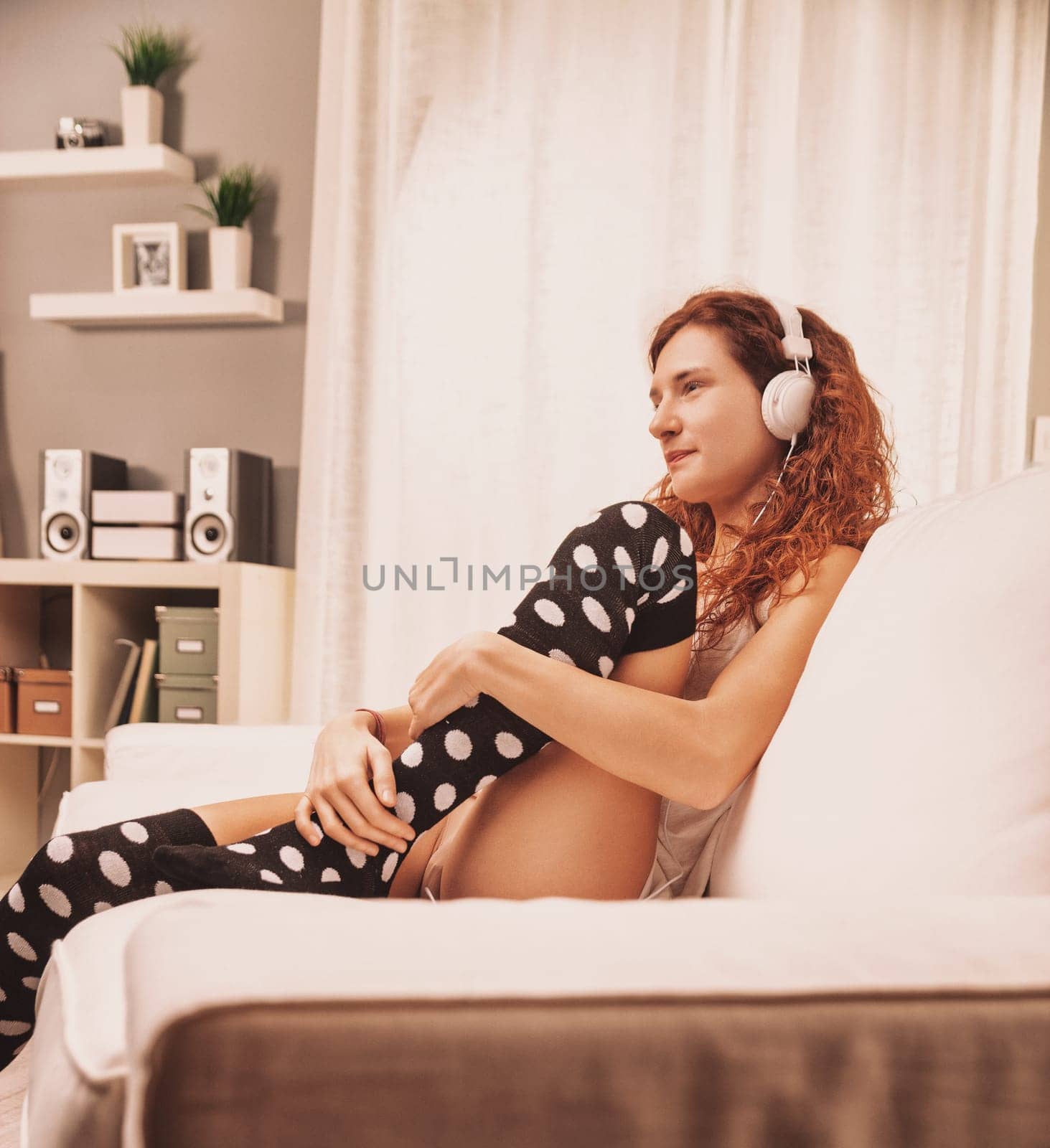 Young woman with long, curly red hair is sitting on a white sofa in her living room, wearing polka dot socks and headphones, enjoying music and looking away