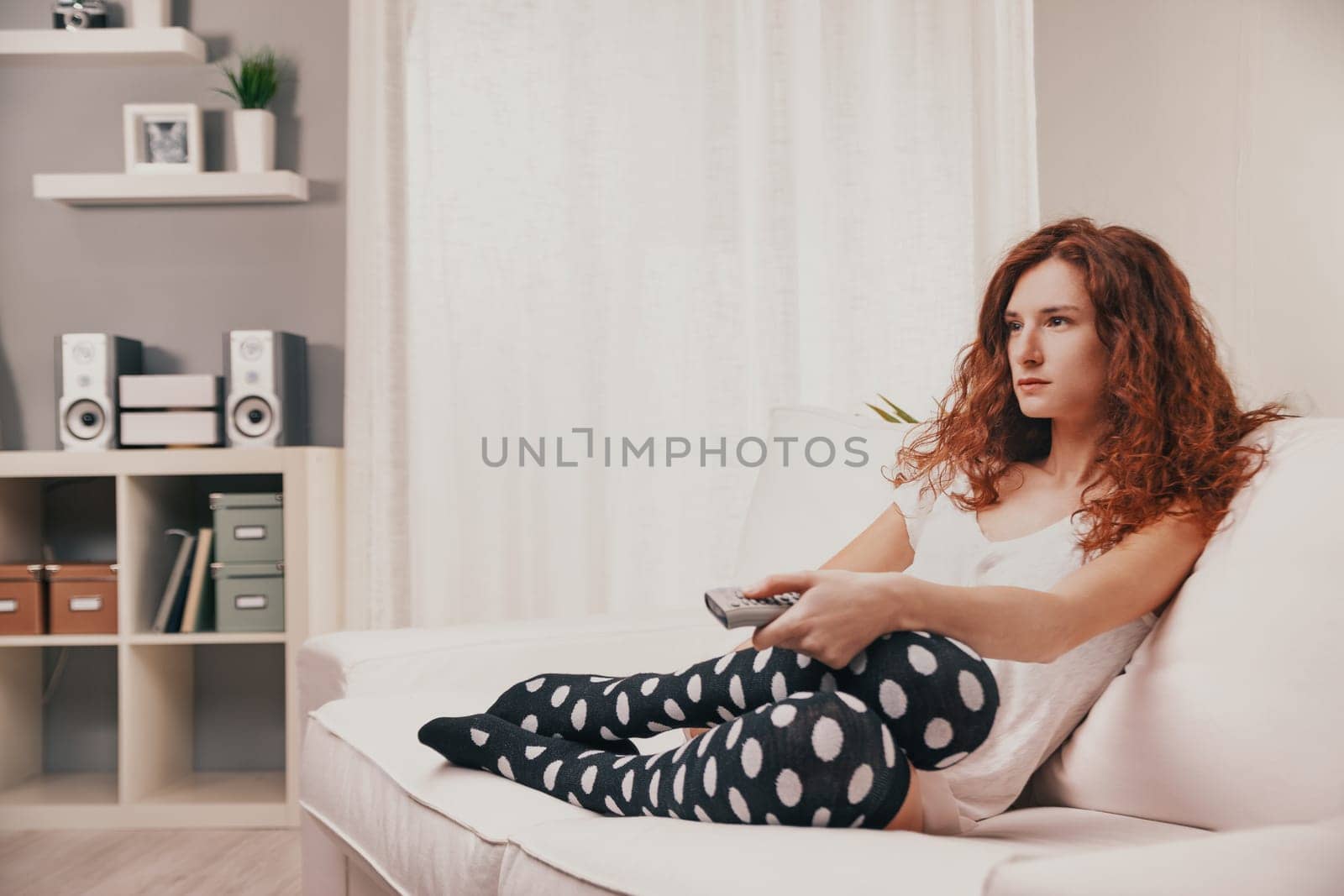Bored young woman with long red curly hair sits on a white sofa in her cozy living room, holding a remote control and watching television, lost in thought and daydreaming