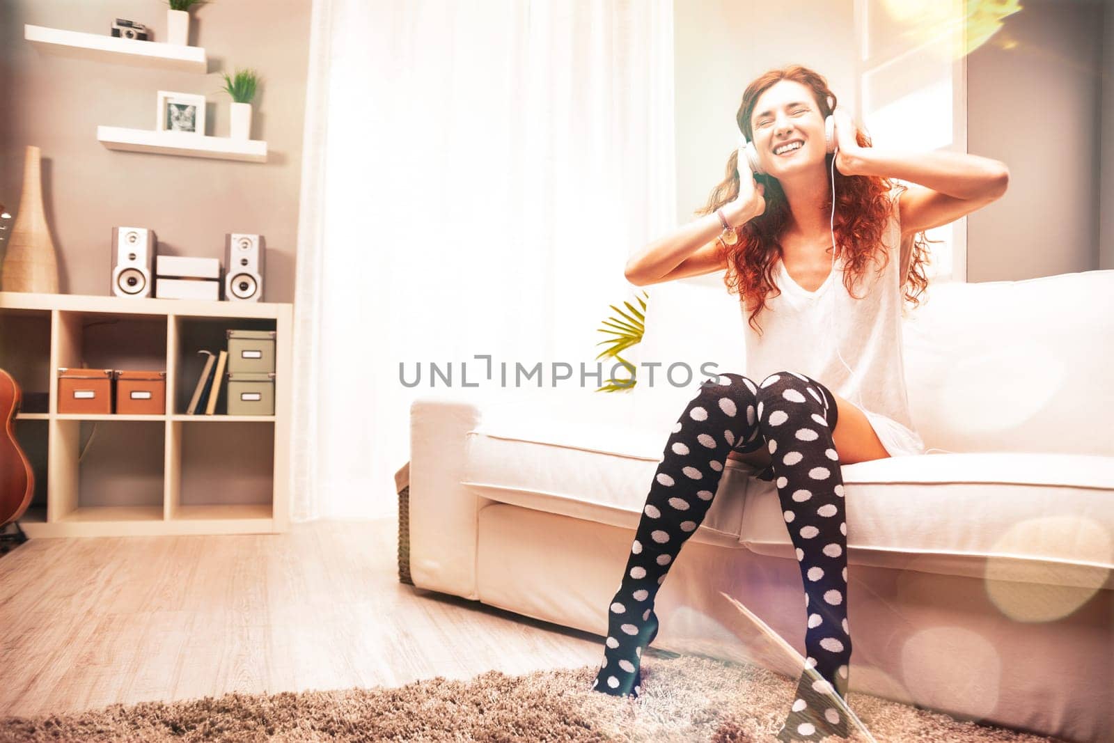Happy young woman sitting comfortably on a white sofa in her modern apartment, enjoying music through headphones, radiating joy and relaxation in her cozy living space