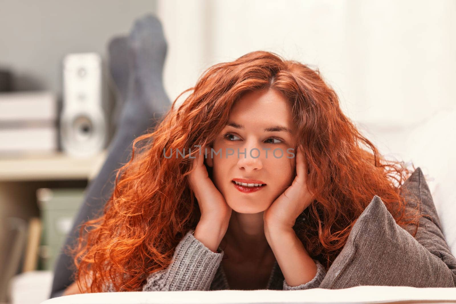 Young woman with long, curly red hair relaxing on a white sofa, resting her head on her hands while enjoying music. Smiling and daydreaming in a cozy living room filled with natural light