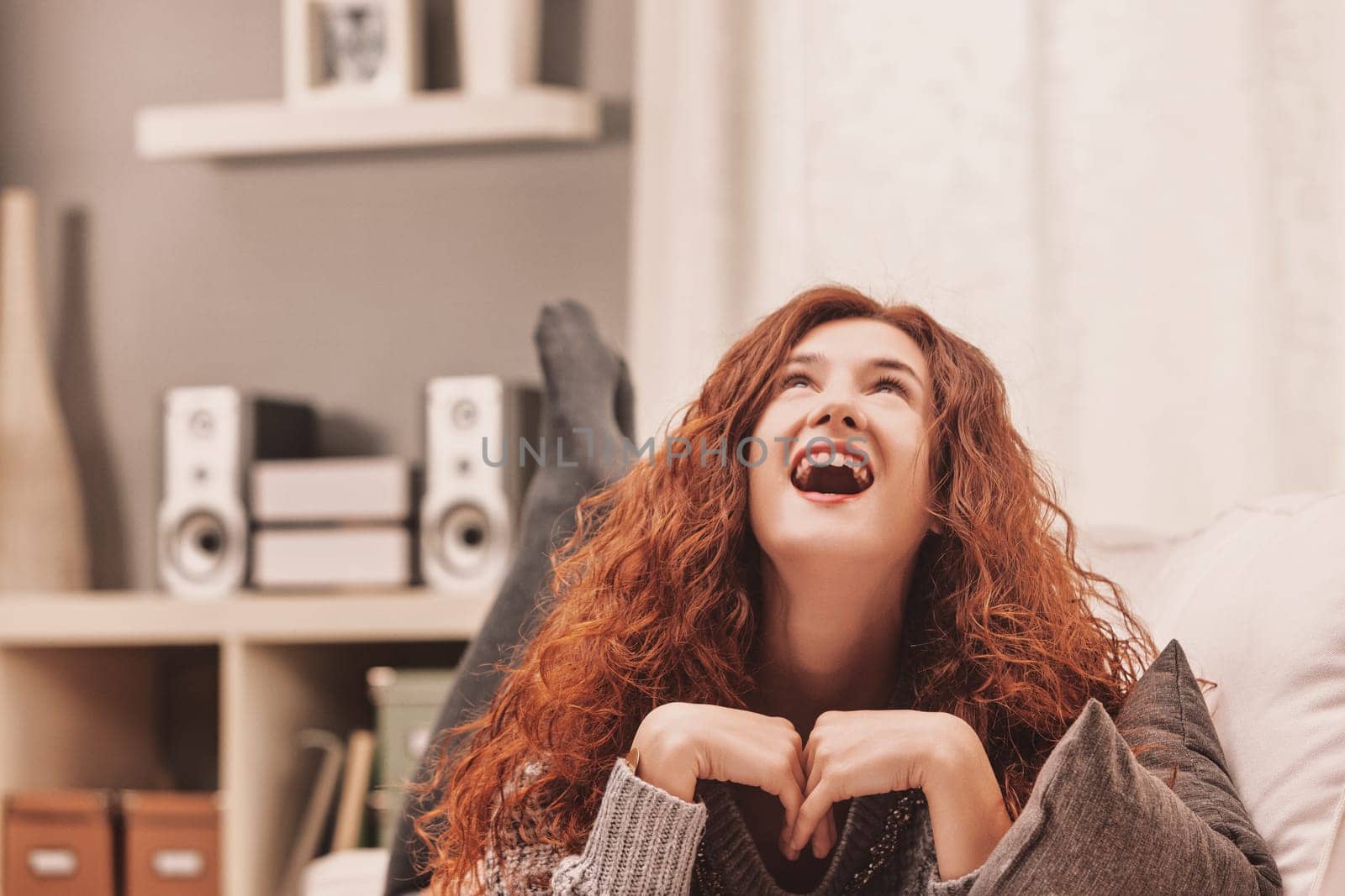 Carefree young woman with long, red curly hair lying on a comfortable sofa with her legs up, laughing and looking up, enjoying her leisure time at home