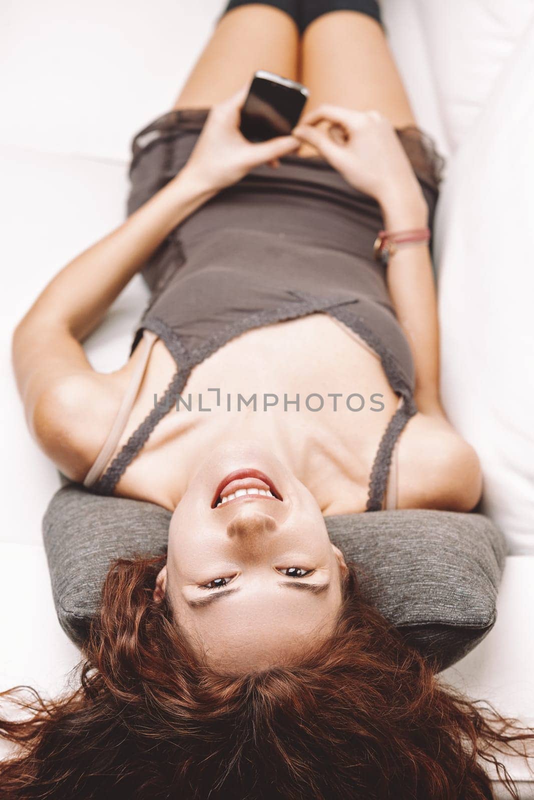 Young woman with long, curly red hair lying upside down on a white sofa, smiling while holding a smartphone, radiating happiness and enjoying a moment of relaxation indoors