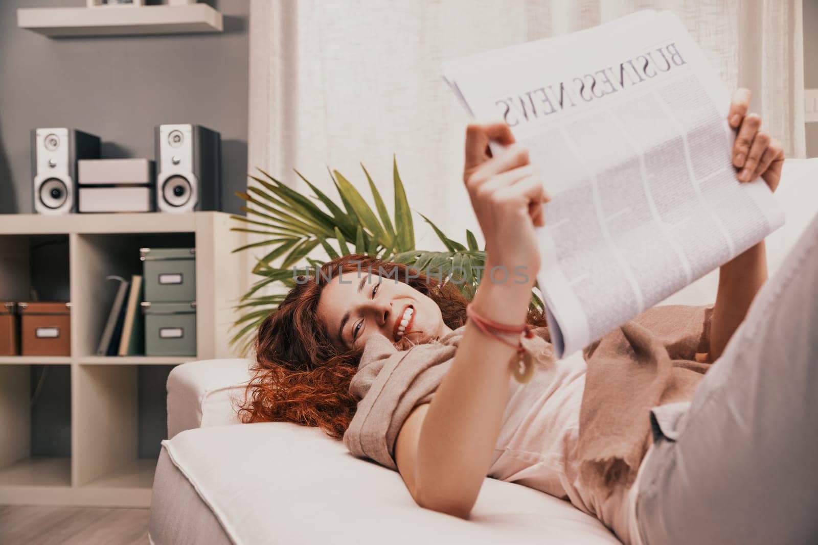 Young woman with long red hair is lying on a white sofa in her living room, reading the business news section of a newspaper and smiling, enjoying her relaxing morning routine