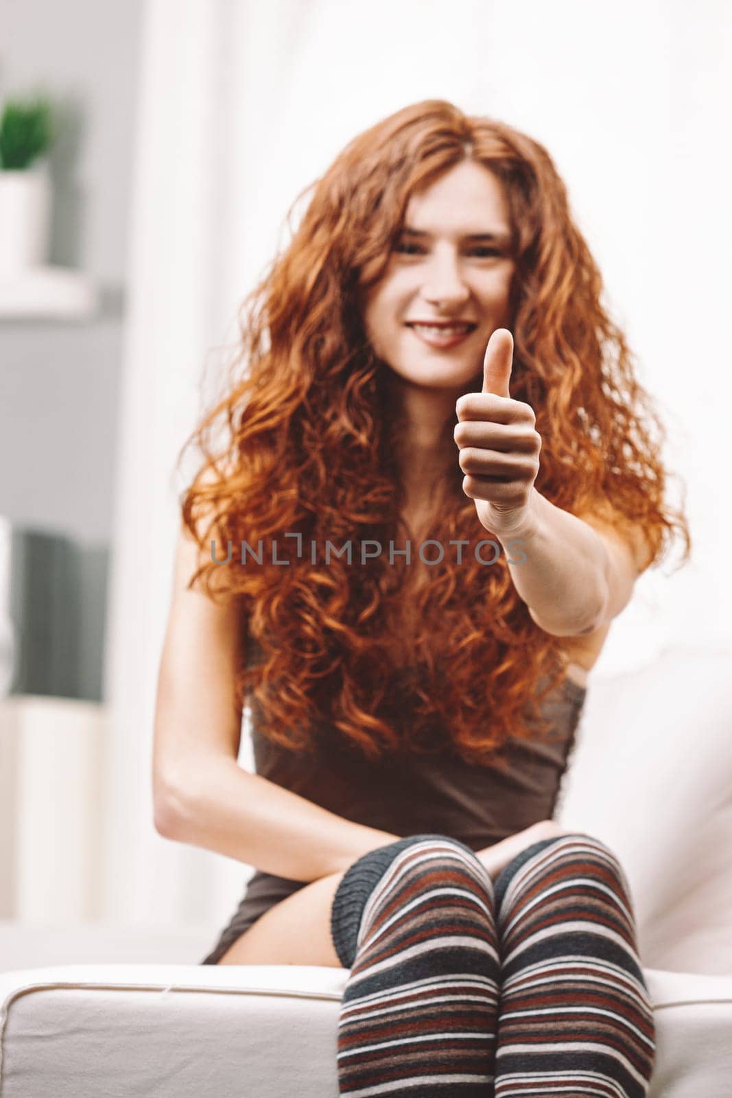 Smiling young woman with long, curly red hair sitting on a white sofa, giving a thumbs up gesture while radiating positivity and confidence, embodying happiness and approval