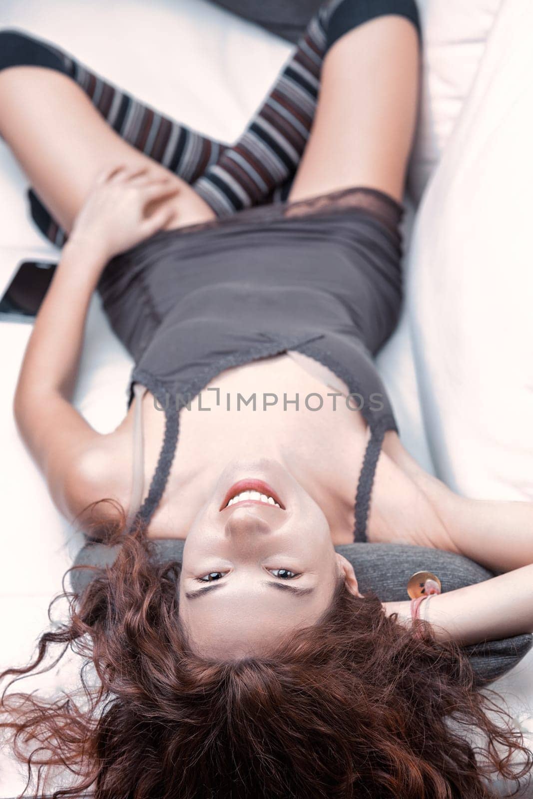Young woman with long, curly red hair smiling while lying upside down on a white sofa, dressed in a stylish gray dress and playful striped socks, radiating joy and relaxation