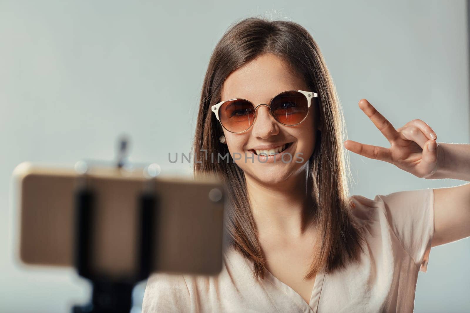 Smiling teenager girl wearing sunglasses is making the peace sign with her fingers while recording a video blog or selfie with her smartphone on a tripod