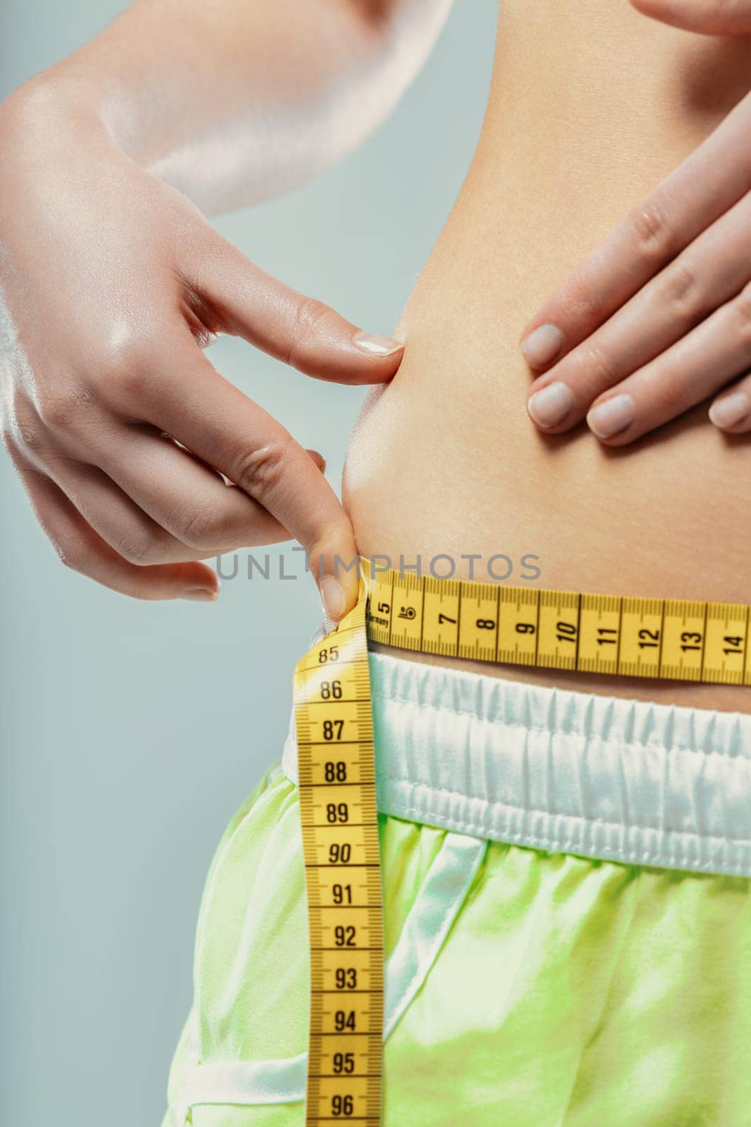 Close-up of a young woman pinching her belly fat while measuring her waist with a yellow measuring tape, expressing concerns about her weight and body image