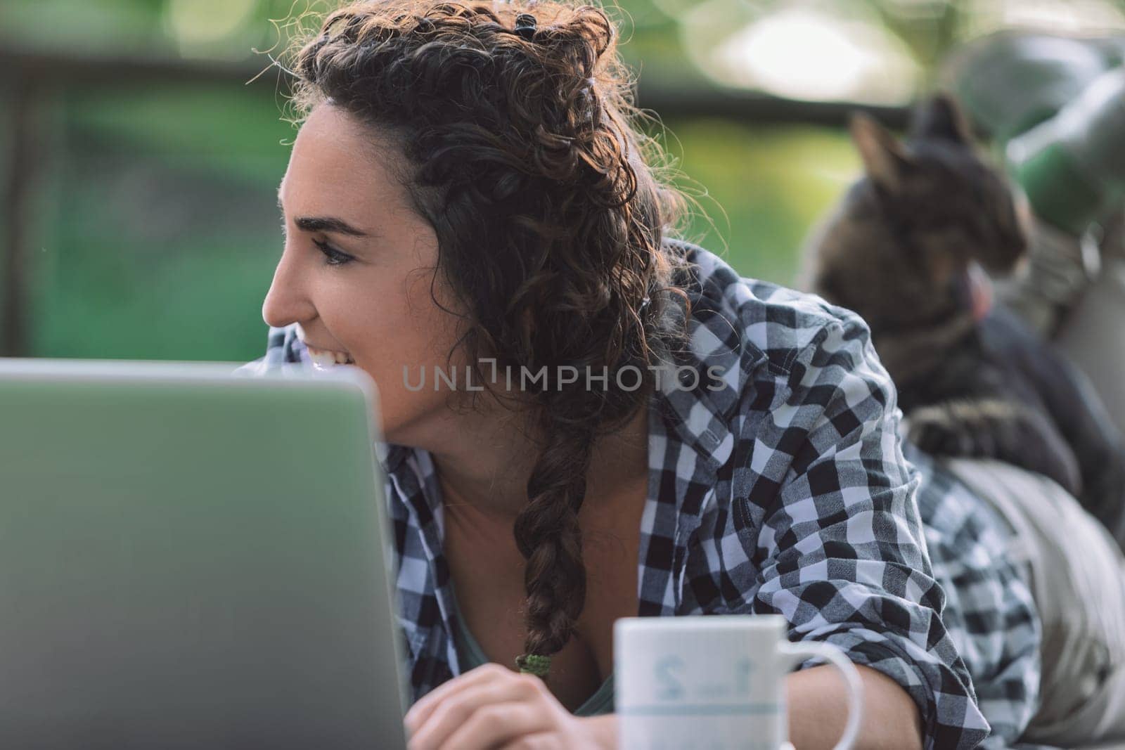 Young woman is lying on her stomach on a balcony, working on her laptop with her cat lying on her back, enjoying the fresh air and sunshine