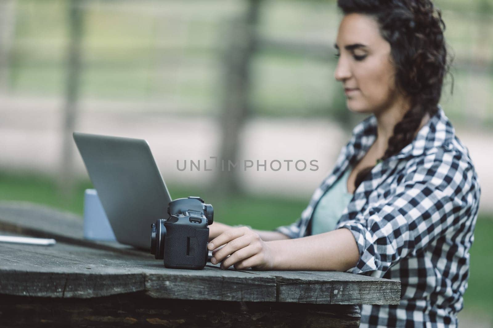 Young adult photographer working on a laptop with her camera on a wooden table in a natural environment, possibly reviewing or editing photos