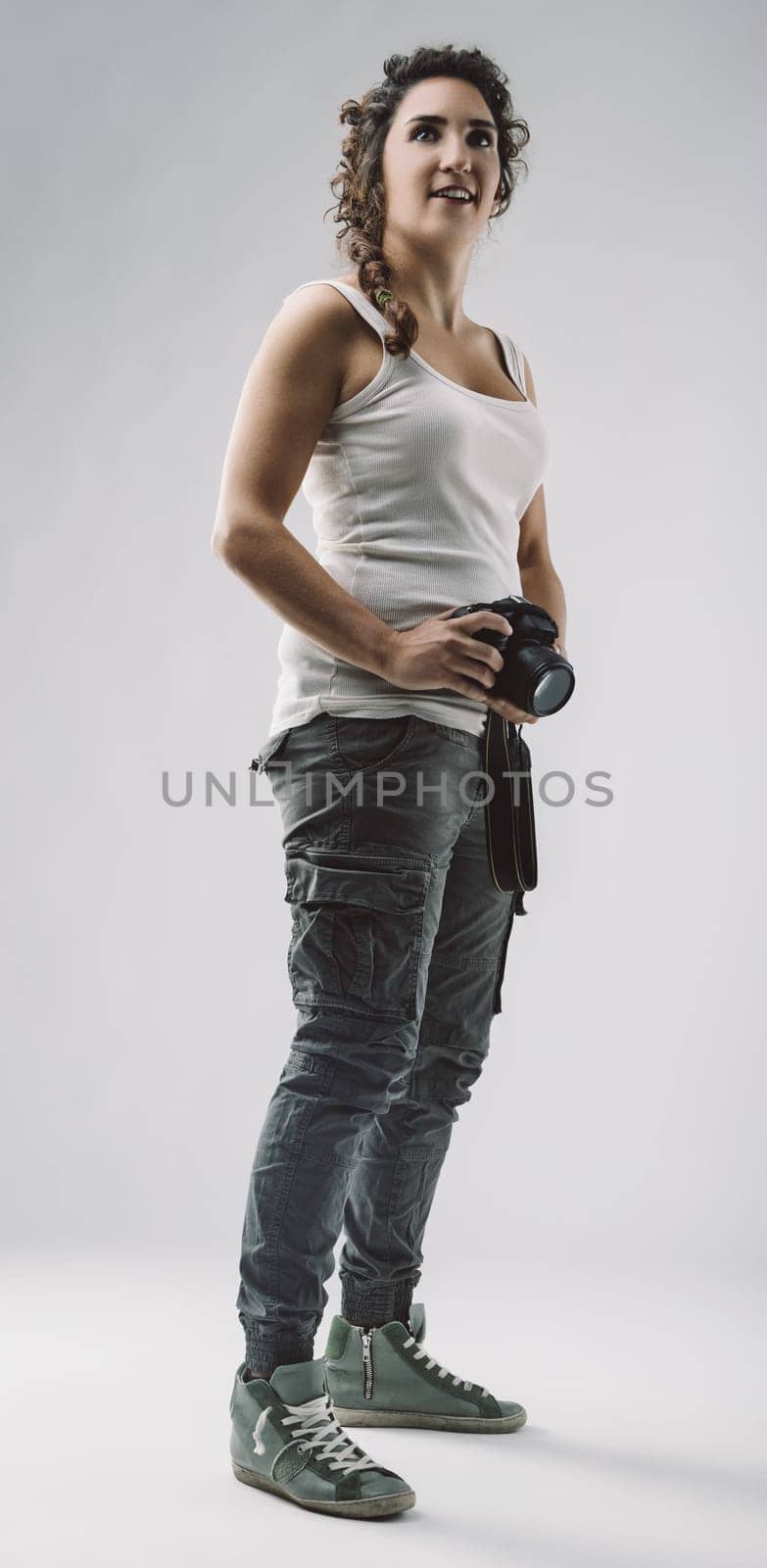 Young female photographer standing in a studio, holding a camera and gazing thoughtfully upwards, embodying creativity and inspiration in her casual tank top and cargo pants