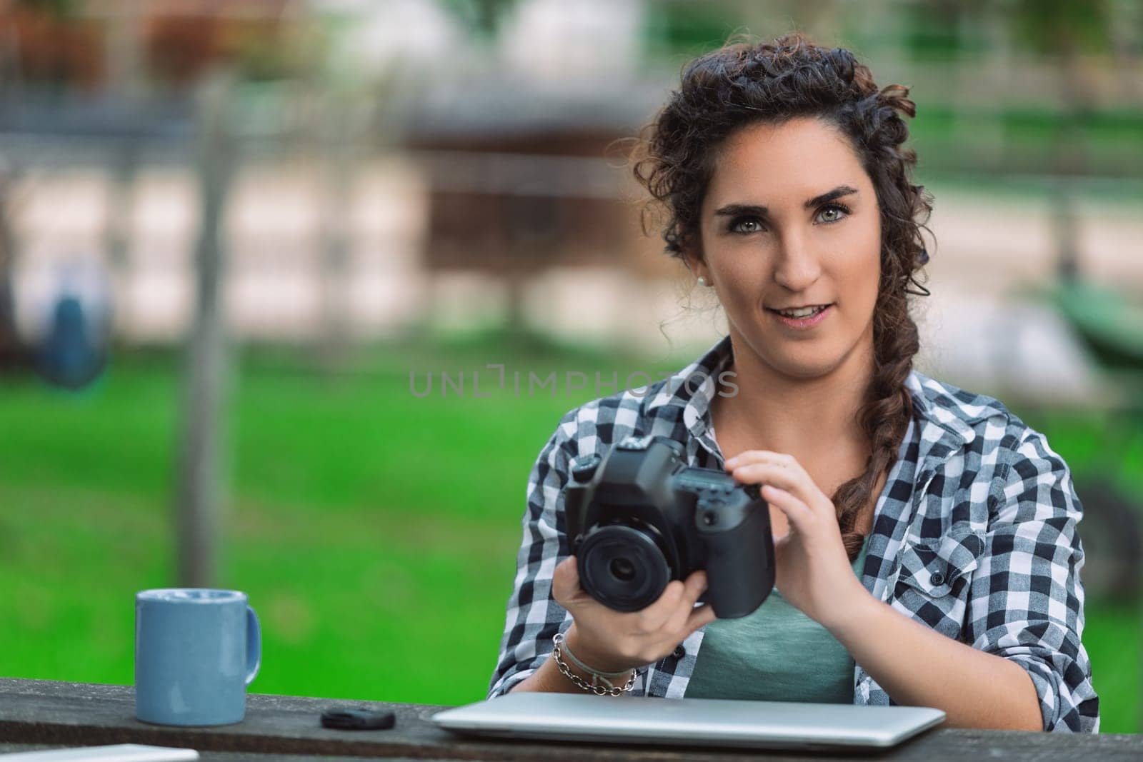 Young female photographer sitting at a table in a park, checking photos on her digital camera while enjoying a cup of coffee and working on her laptop amidst nature's greenery