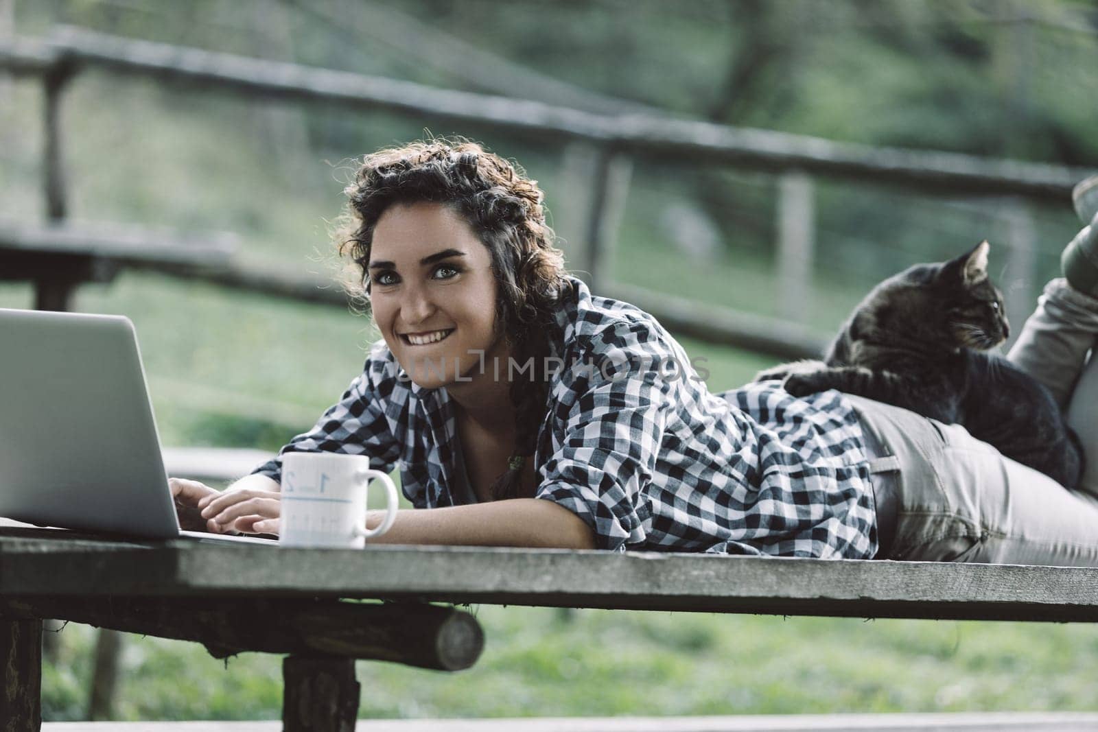 Young woman lying on a wooden picnic table, working on her laptop while sipping coffee. A cat comfortably resting on her back adds a cozy touch to the serene outdoor scene