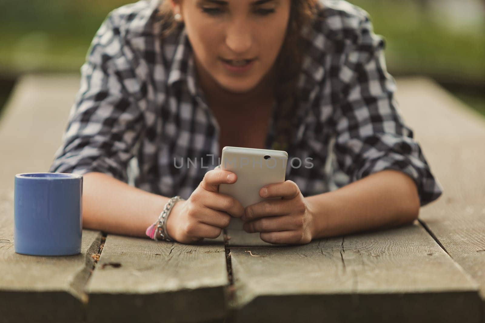 Young woman lounging on a picnic table, using her mobile phone while a mug sits beside her, savoring leisure moments outdoors in the warm summer sunlight