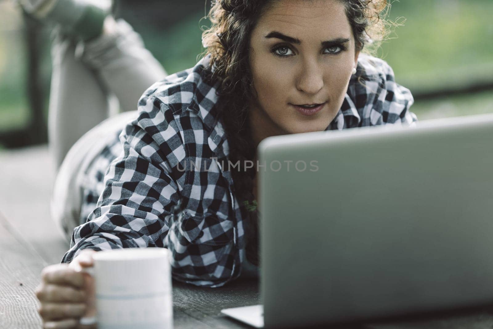Young woman lying on a wooden table outdoors, working on her laptop while holding a mug of coffee, smiling confidently and looking directly at the camera in a relaxed, casual setting
