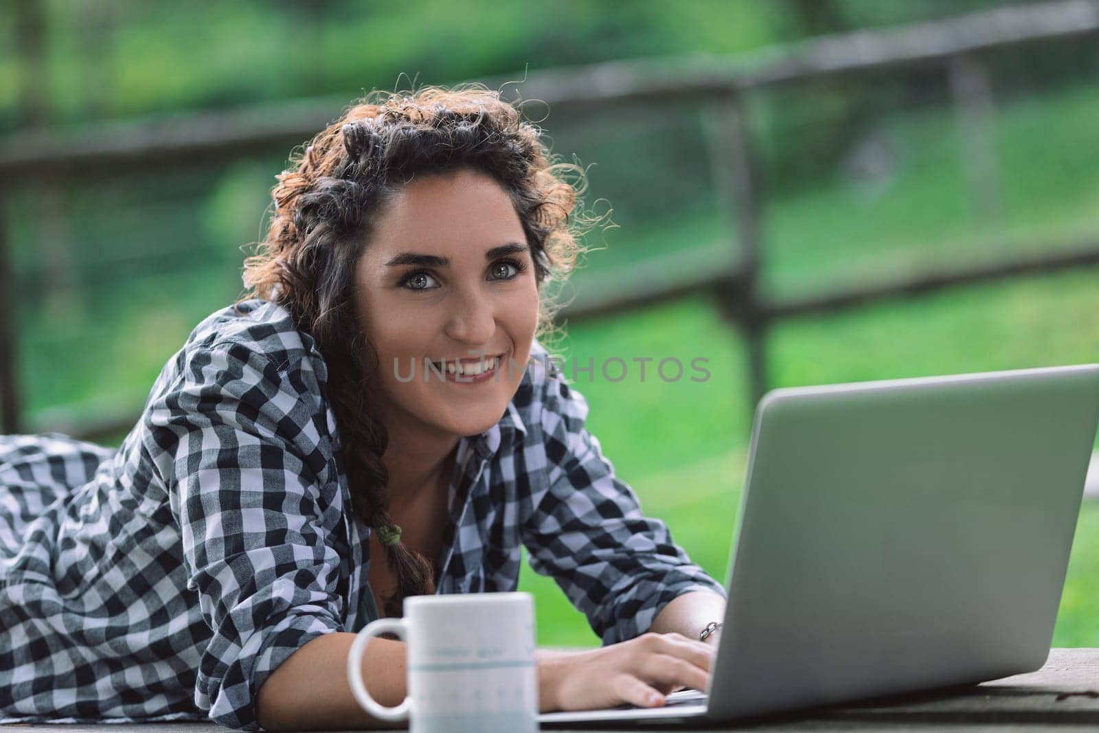 Young woman is lying on her stomach on a picnic table, working on her laptop with a mug next to her, enjoying the fresh air and greenery of a park