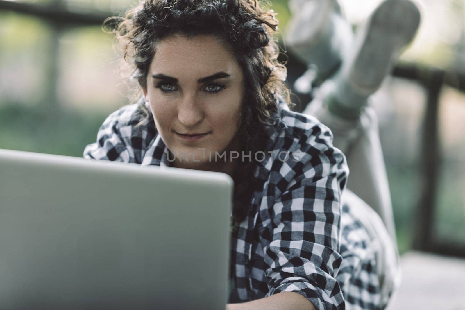 Young woman with curly hair, wearing a checkered shirt, is lying down on a deck or patio, looking intently at her laptop and working outdoors