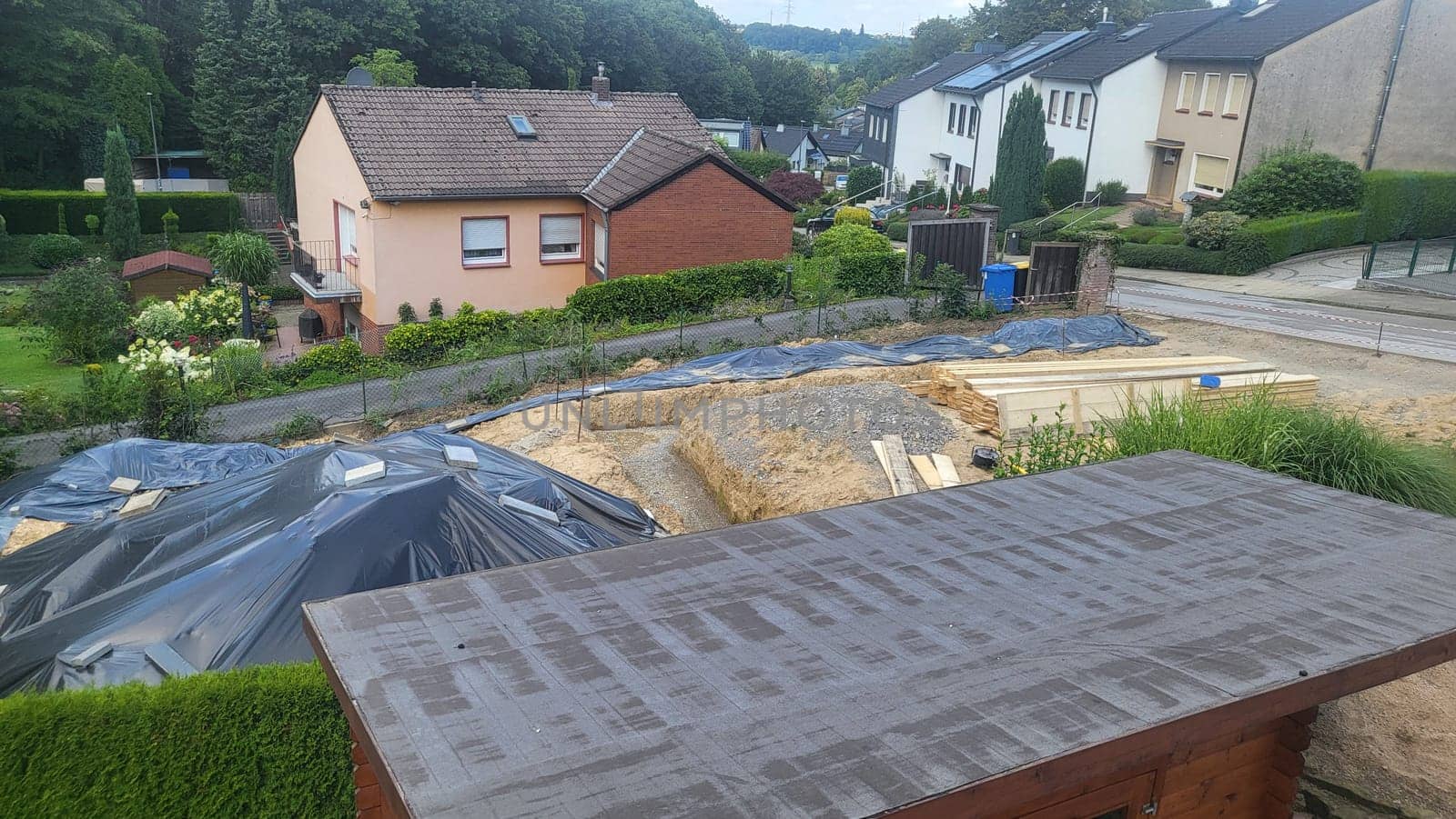 A construction site in a suburban neighborhood, featuring an excavated area covered with a tarp, a wooden shed, and residential houses in the background.