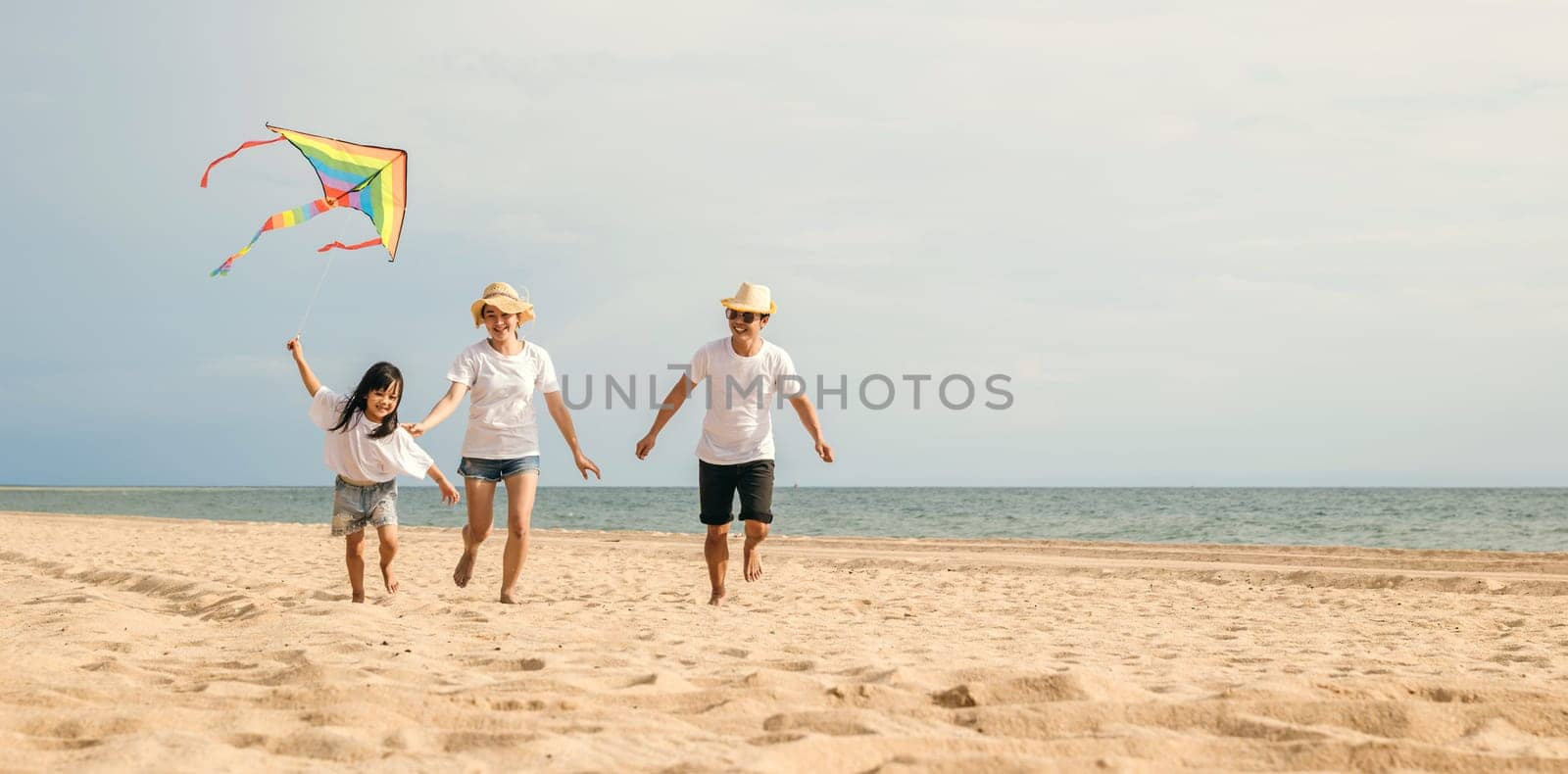 Asian family is flying a kite high in the sky while running on the beach. Happy parents mother and father with their child playing with kite, family is enjoying their time together, summer day