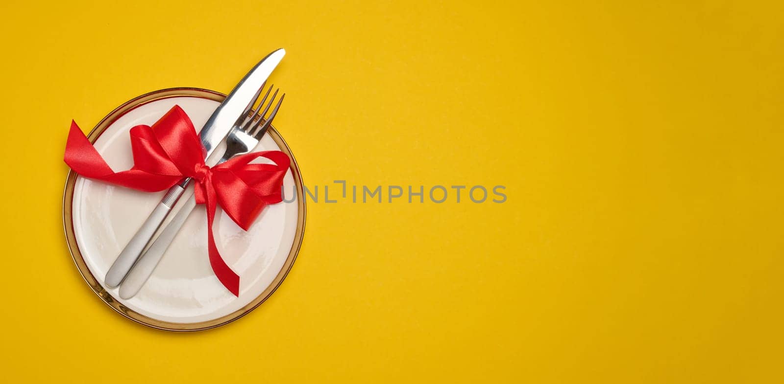 Round white plate and tied with red bow view with knife on yellow background, festive table setting. Copy space