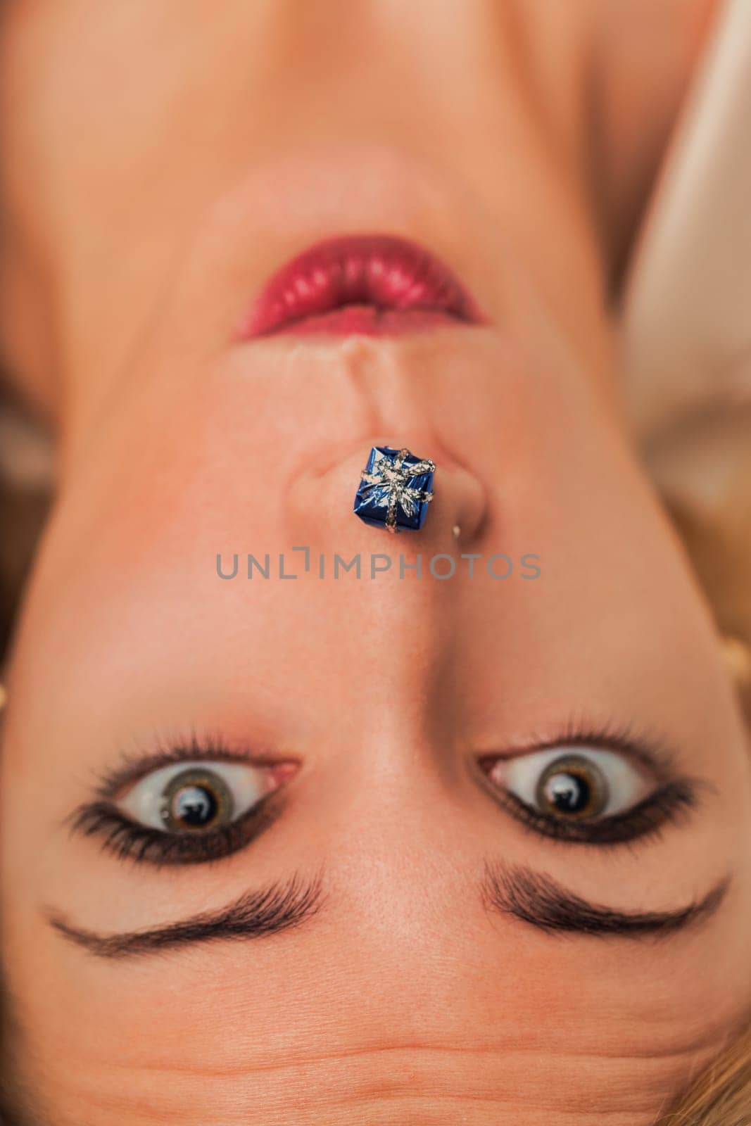Young woman lying down with a small blue gift placed under her nose, expressing surprise and curiosity in a playful and intriguing composition
