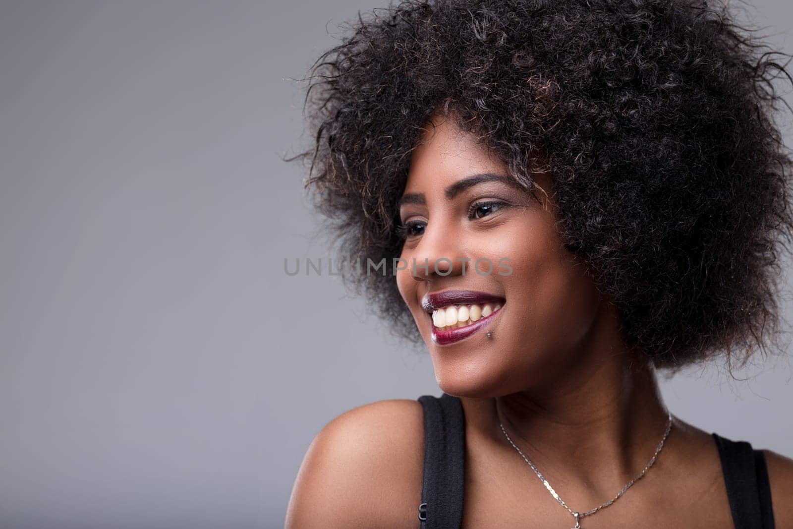 Studio portrait of a beautiful young black woman with curly hair, smiling and looking away on a gray background, expressing happiness and positivity